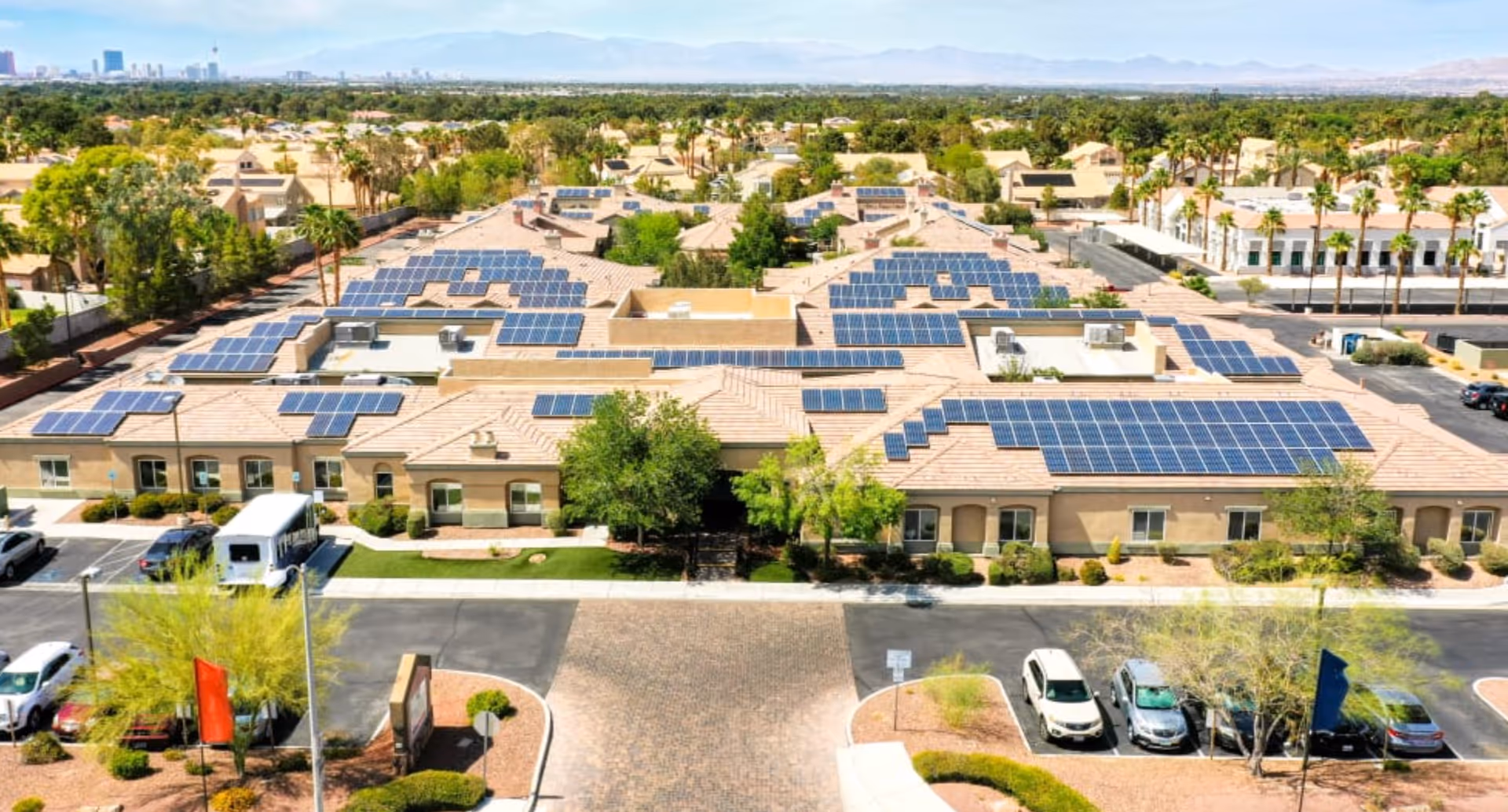 Aerial view of a senior living facility named Cottages at Green Valley, showing multiple single-story buildings with solar panels on the roofs, surrounded by parking lots, trees, and landscaped areas. The city skyline and mountains are visible in the background under a clear sky.