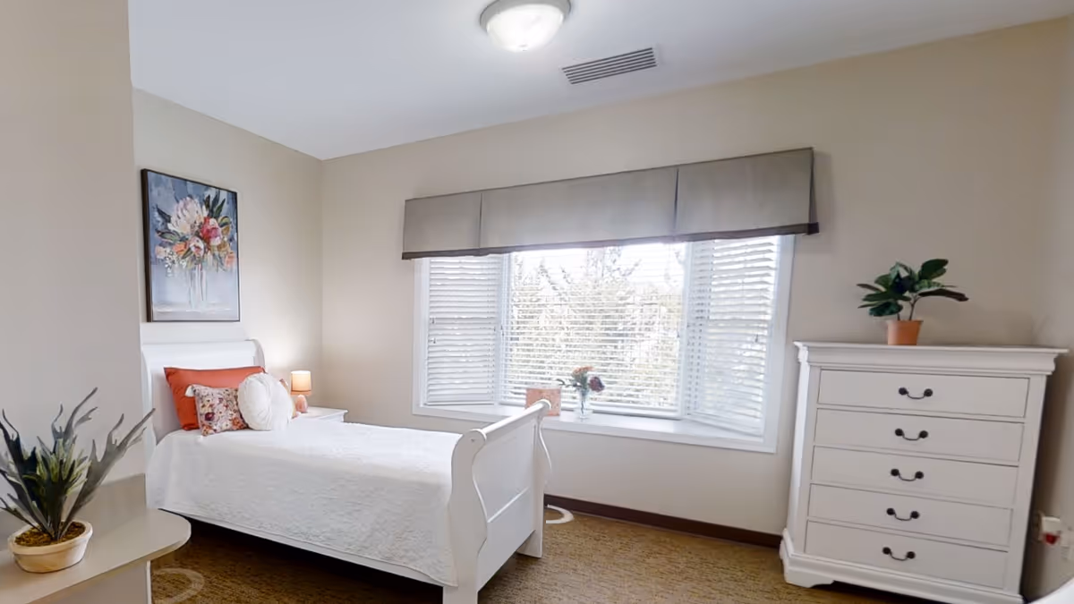A bright bedroom with a white bed featuring orange and floral pillows, a white nightstand with a small lamp, a large window with blinds and a valance, a white dresser with a potted plant on top, and a floral painting on the wall.