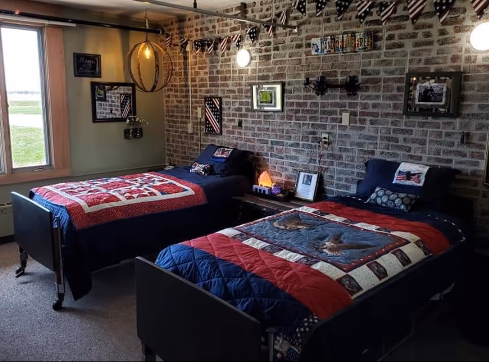 A cozy bedroom with two single beds featuring patriotic-themed quilts and pillows. The room has a brick accent wall decorated with framed pictures, a string of small American flags hanging from the ceiling, and a window letting in natural light. There is a wooden nightstand between the beds with a lamp and framed photo.