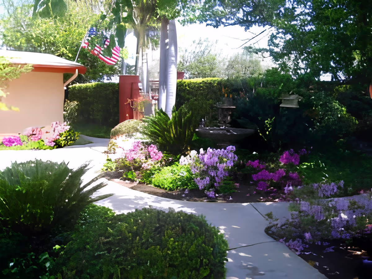 A garden area with a concrete pathway winding through lush green bushes and colorful flowering plants. There are tall trees and a small building with a red roof on the left side. An American flag is visible near the building, and the scene is brightly lit by sunlight.
