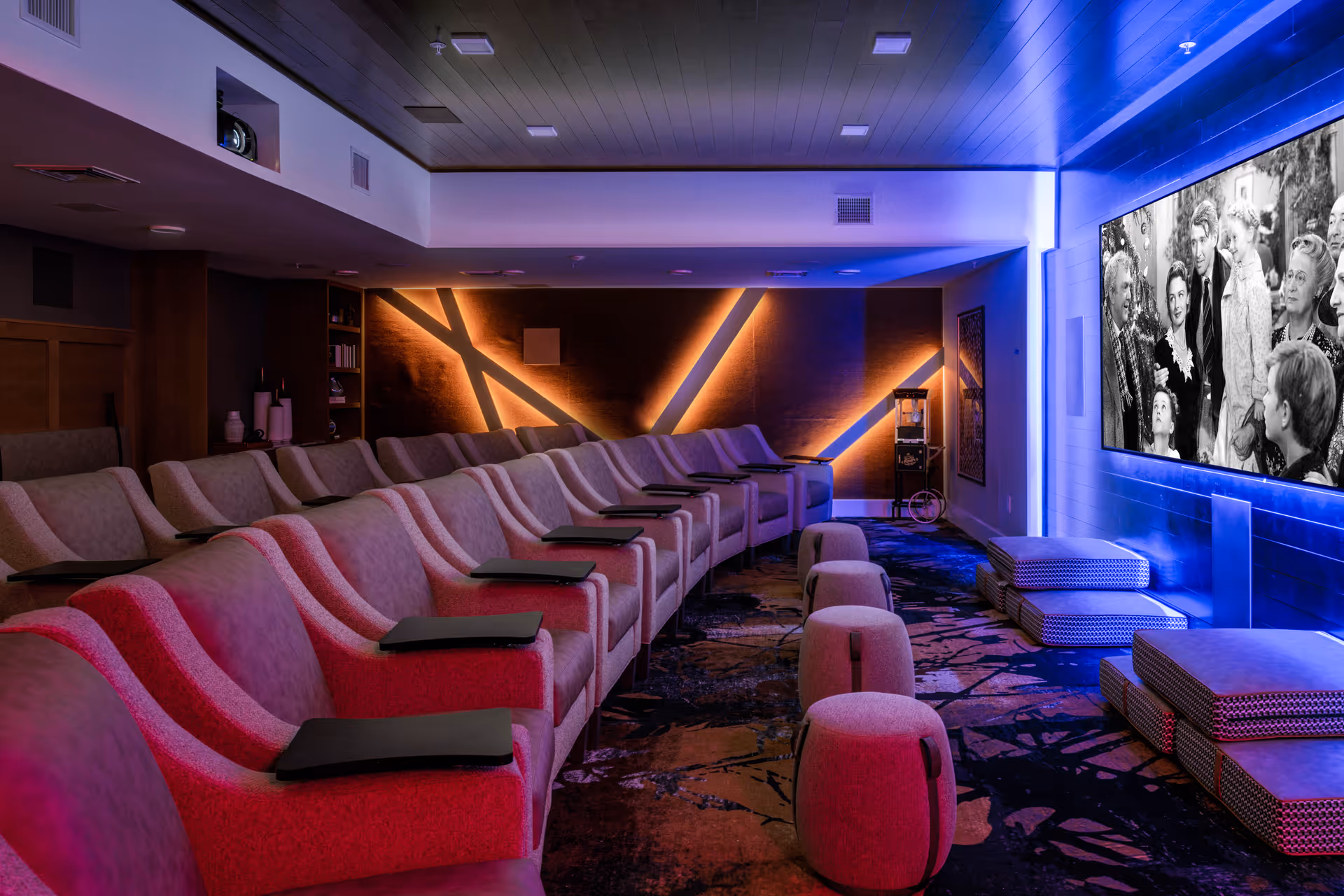 A modern home theater room with multiple rows of comfortable armchairs equipped with small black trays. The room features ambient lighting with orange and blue hues, a large screen displaying a black and white movie scene, and several cushioned stools and ottomans in front of the screen.