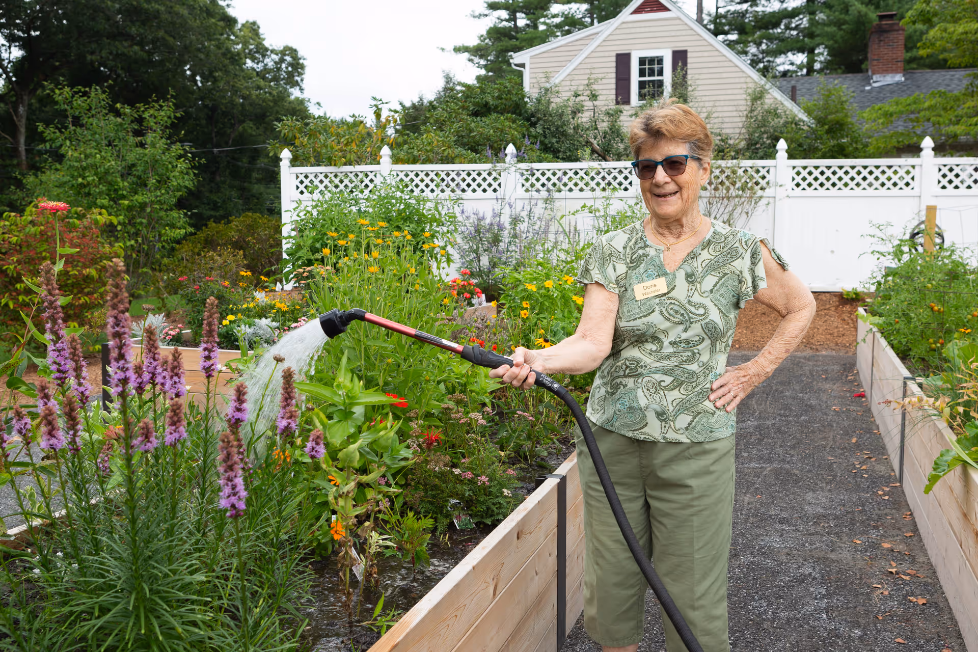 An elderly woman wearing sunglasses and a green patterned shirt waters plants in a raised garden bed using a garden hose. The garden is lush with various flowers and greenery, and there is a white lattice fence and a house in the background.