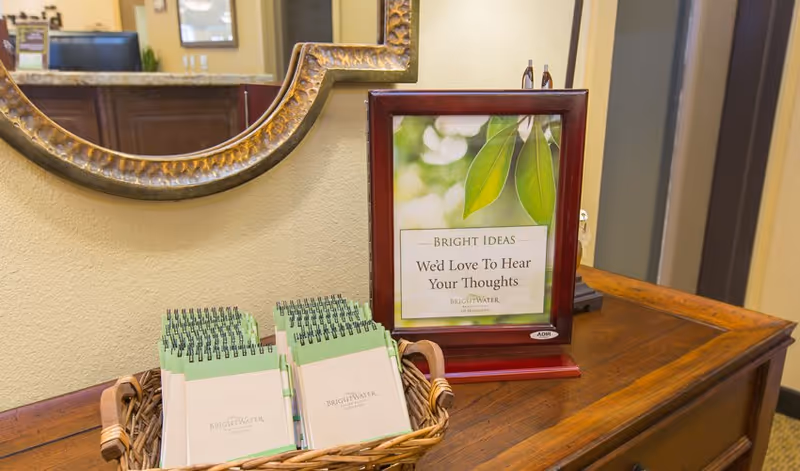 A wooden table with a basket holding several small spiral notebooks. Next to the basket is a framed sign with a green leaf image and text that reads 'Bright Ideas We'd Love To Hear Your Thoughts Brightwater Senior Living of Highland.' A decorative mirror hangs on the wall above the table, and a reception desk is visible in the background.
