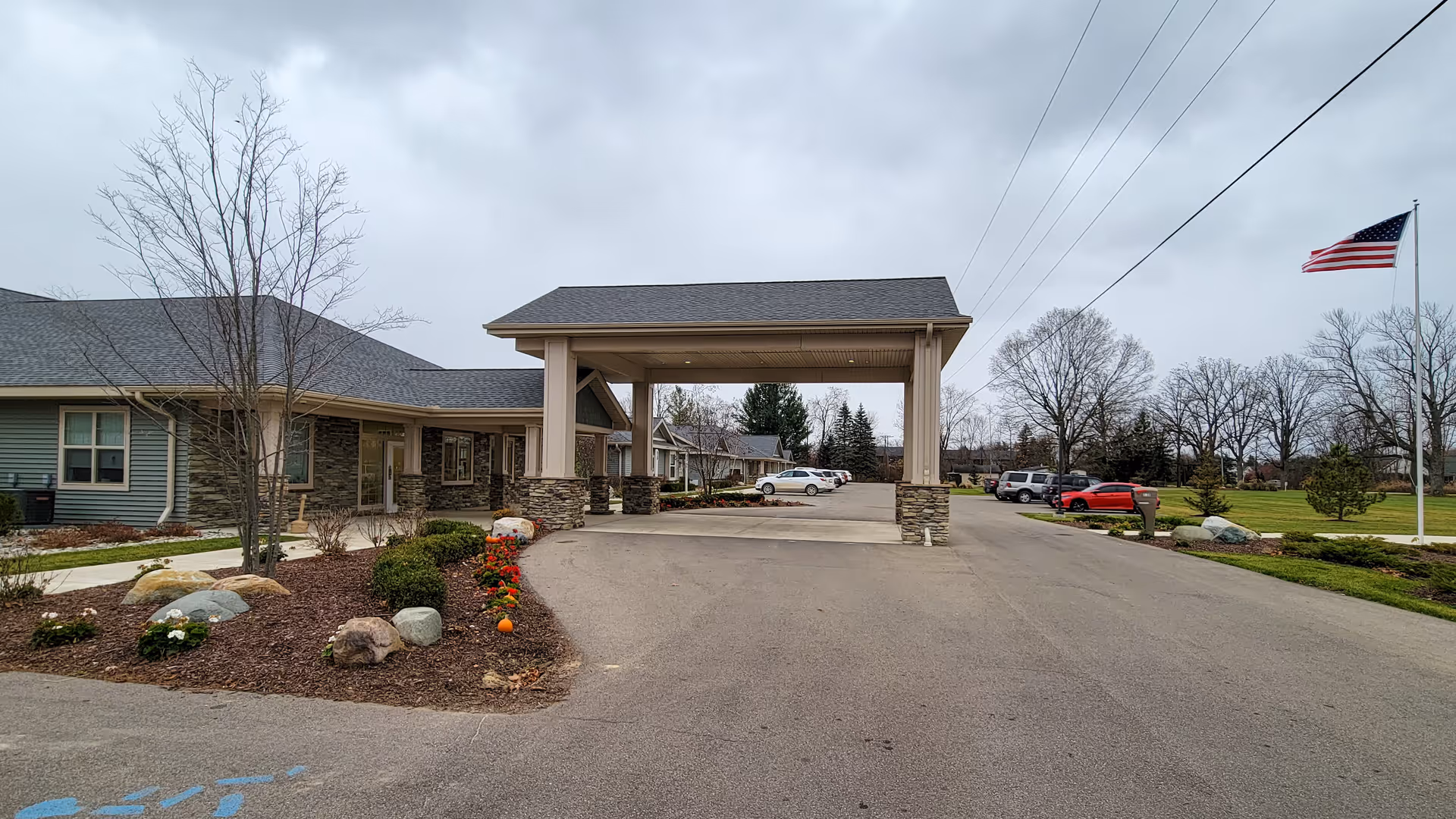 Front entrance and porte-cochere of a single-story senior living facility with landscaped grounds, parked cars, and an American flag.