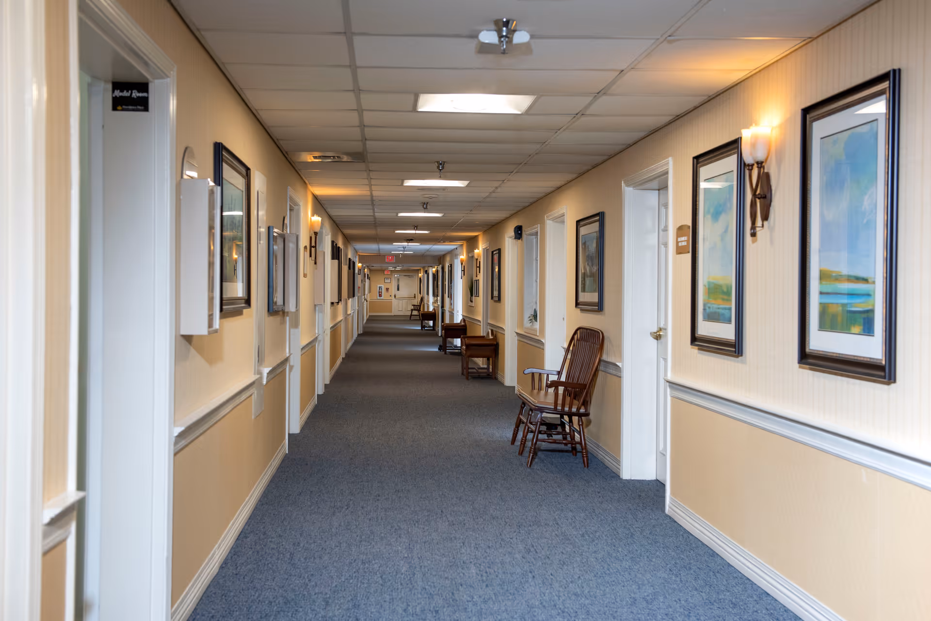 A long, well-lit hallway in a senior living facility with beige walls, blue carpet, framed artwork, and wooden chairs placed along the walls. Several white doors line both sides of the corridor.