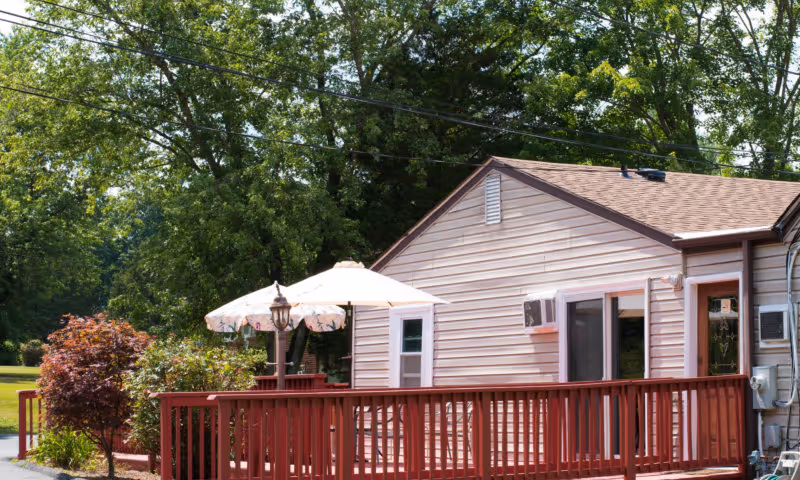 Outdoor view of a single-story building with beige siding and a brown roof, featuring a red wooden deck with a railing. On the deck, there are two white patio umbrellas and some greenery including bushes and trees in the background.