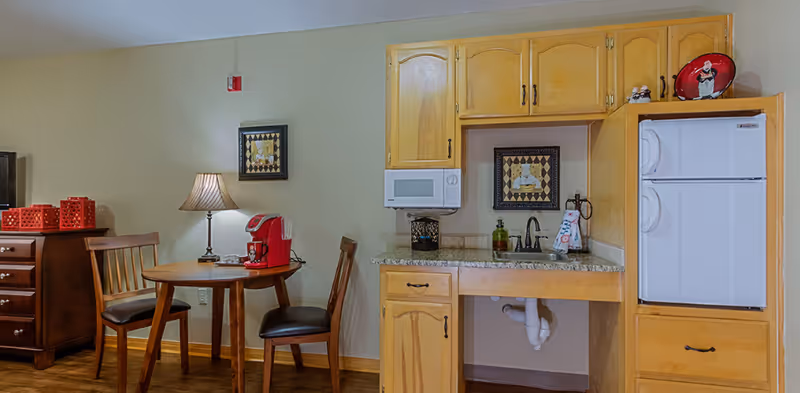 Small kitchenette area with wooden cabinets, a white microwave, and a white refrigerator. There is a granite countertop with a sink and soap dispenser. Next to the kitchenette is a round wooden table with two wooden chairs, a table lamp, and a red coffee maker. The walls are beige with framed artwork hanging above the sink and table.