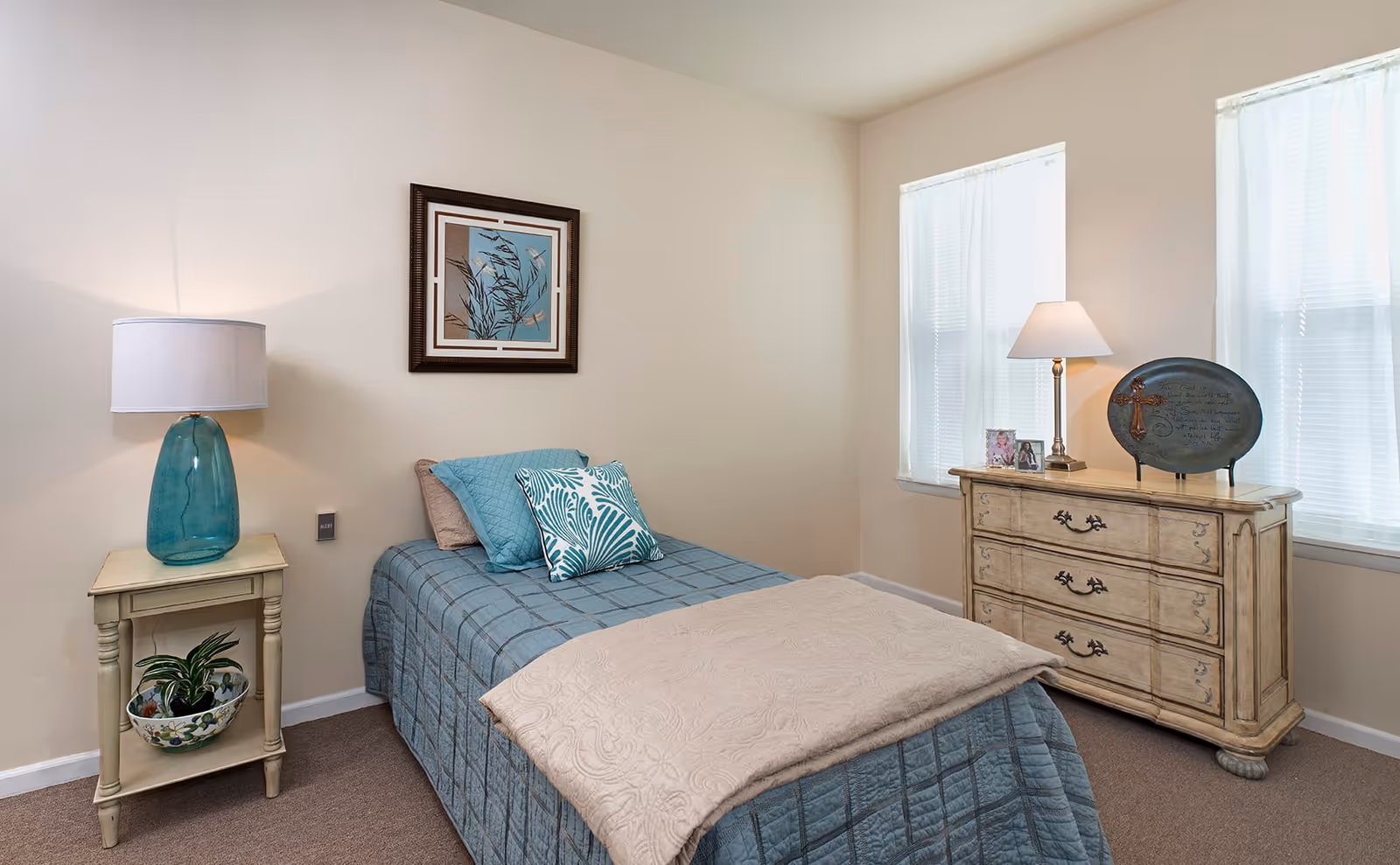 A cozy bedroom with a single bed covered in blue and beige bedding, a small wooden nightstand with a blue glass lamp and a potted plant, a wooden dresser with a lamp, framed photos, and a decorative plate, and two windows with white curtains letting in natural light.