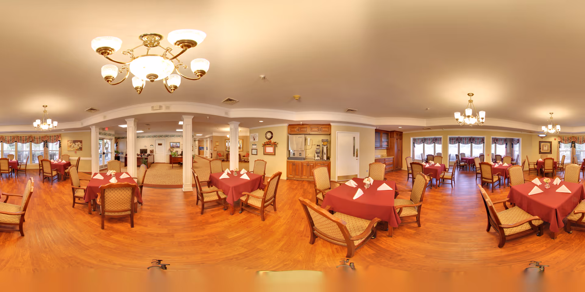 A spacious dining room with multiple tables covered in red tablecloths, each set with folded white napkins. The room features wooden flooring, upholstered chairs, large windows with valances allowing natural light, and elegant chandeliers hanging from the ceiling. The area appears clean and well-organized, suitable for communal dining in a senior living facility.
