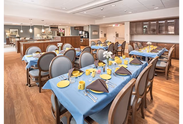A bright and spacious dining room in a senior living facility with multiple tables covered in blue tablecloths, set with yellow plates, mugs, and brown napkins. The room features wooden flooring, comfortable chairs, and a serving area in the background with cabinetry and a coffee station.
