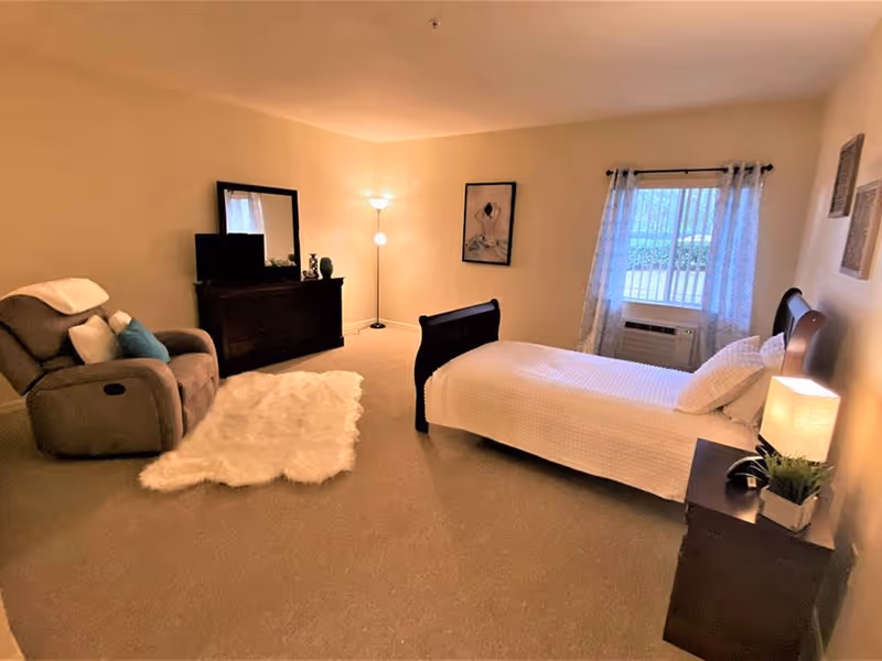 A cozy bedroom with a single bed covered in white bedding, a wooden nightstand with a lamp and a small plant, a recliner chair with pillows and a blanket, a fluffy white rug on the carpeted floor, a dresser with a mirror and a TV on top, a floor lamp in the corner, and a window with sheer curtains letting in natural light.