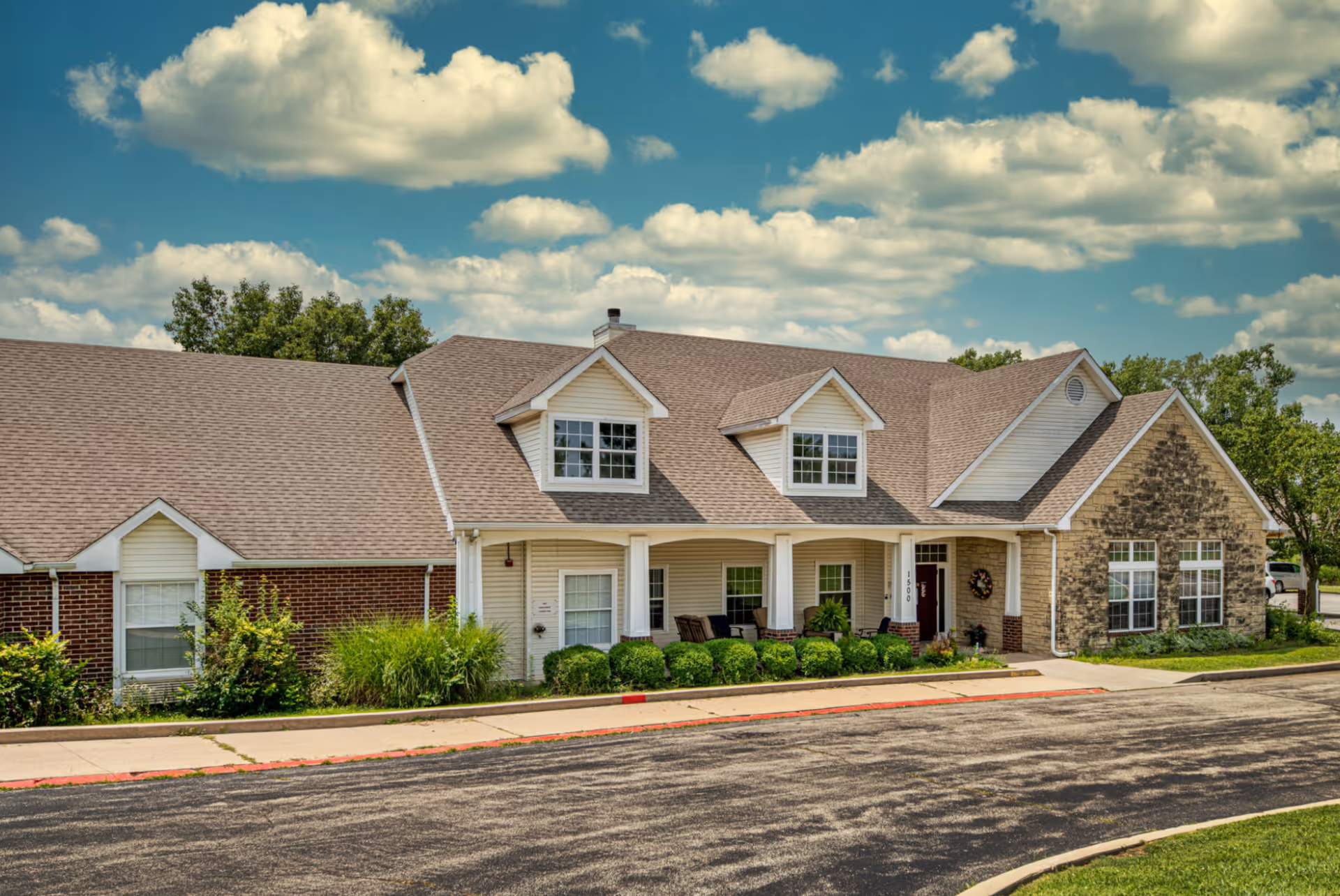 Exterior view of Creekside Village, a single-story building with a combination of brick and stone facade, multiple windows, a covered porch with seating, and a well-maintained lawn under a partly cloudy sky.