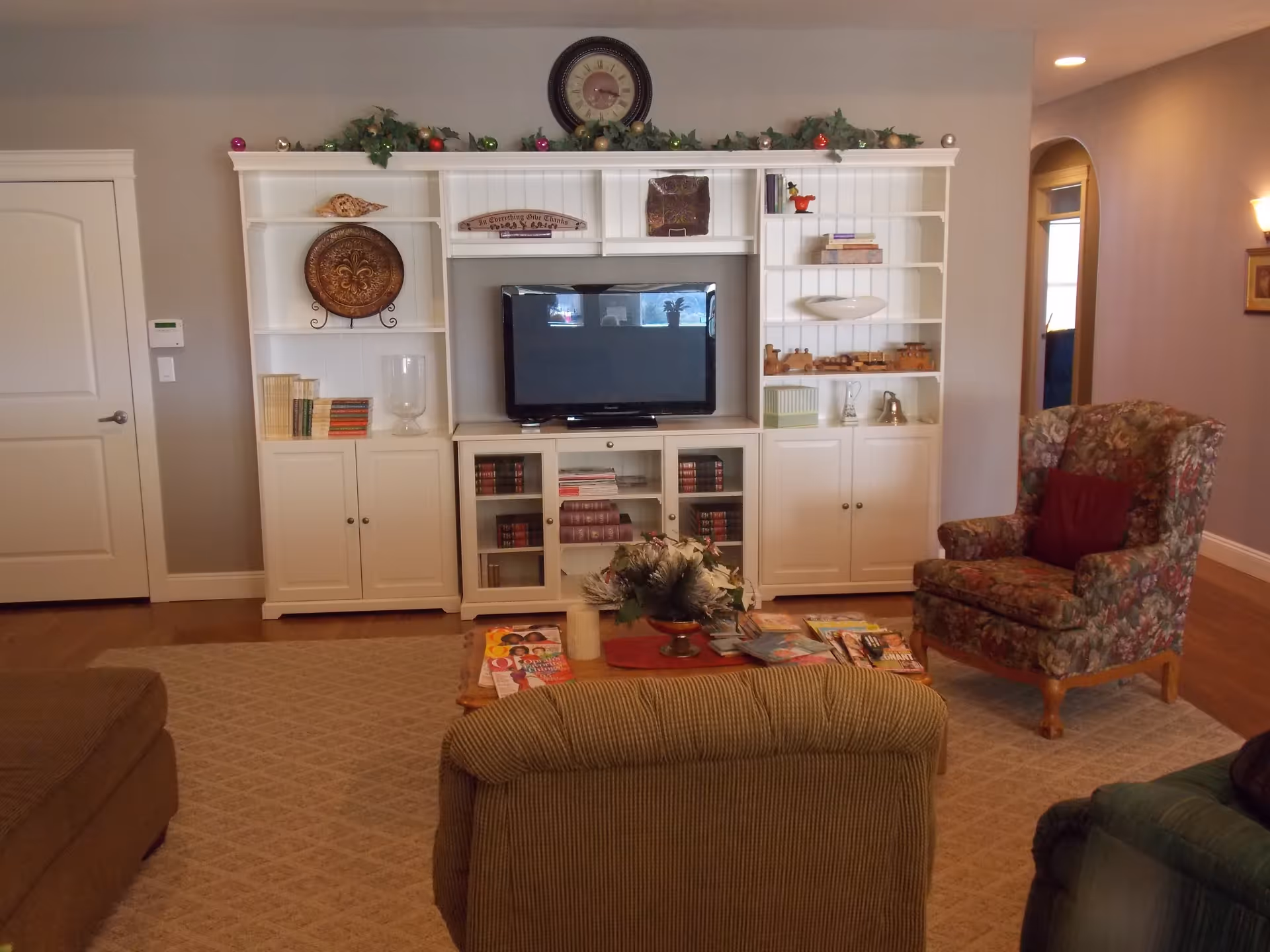 A cozy living room with a white entertainment center holding a flat-screen TV, books, and decorative items. There is a floral upholstered armchair with a red cushion, a green armchair, and a beige armchair surrounding a wooden coffee table with magazines and a floral centerpiece. The room has beige walls, a carpeted floor, and a door on the left side.