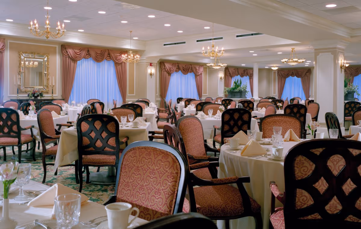 Large elegant dining room with round tables set with white tablecloths, folded napkins, glassware, and upholstered chairs.