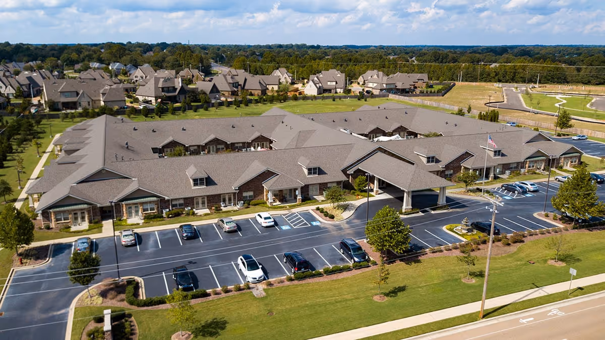 Aerial view of Hearthside Senior Living facility showing a large single-story building with a gray roof, surrounded by a parking lot with several cars parked. The building has a covered entrance with an American flag nearby. The surrounding area includes green lawns, trees, and neighboring residential houses in the background.