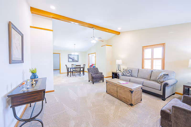 A bright and spacious living room area with beige walls and carpeted floor. The room features a gray sofa with patterned cushions, two gray armchairs, a wooden coffee table, and a side table with a lamp. In the background, there is a dining area with a wooden dining table and chairs, a ceiling fan with lights, and a framed picture on the wall. A small console table with decorative items and a framed picture hangs on the left wall.