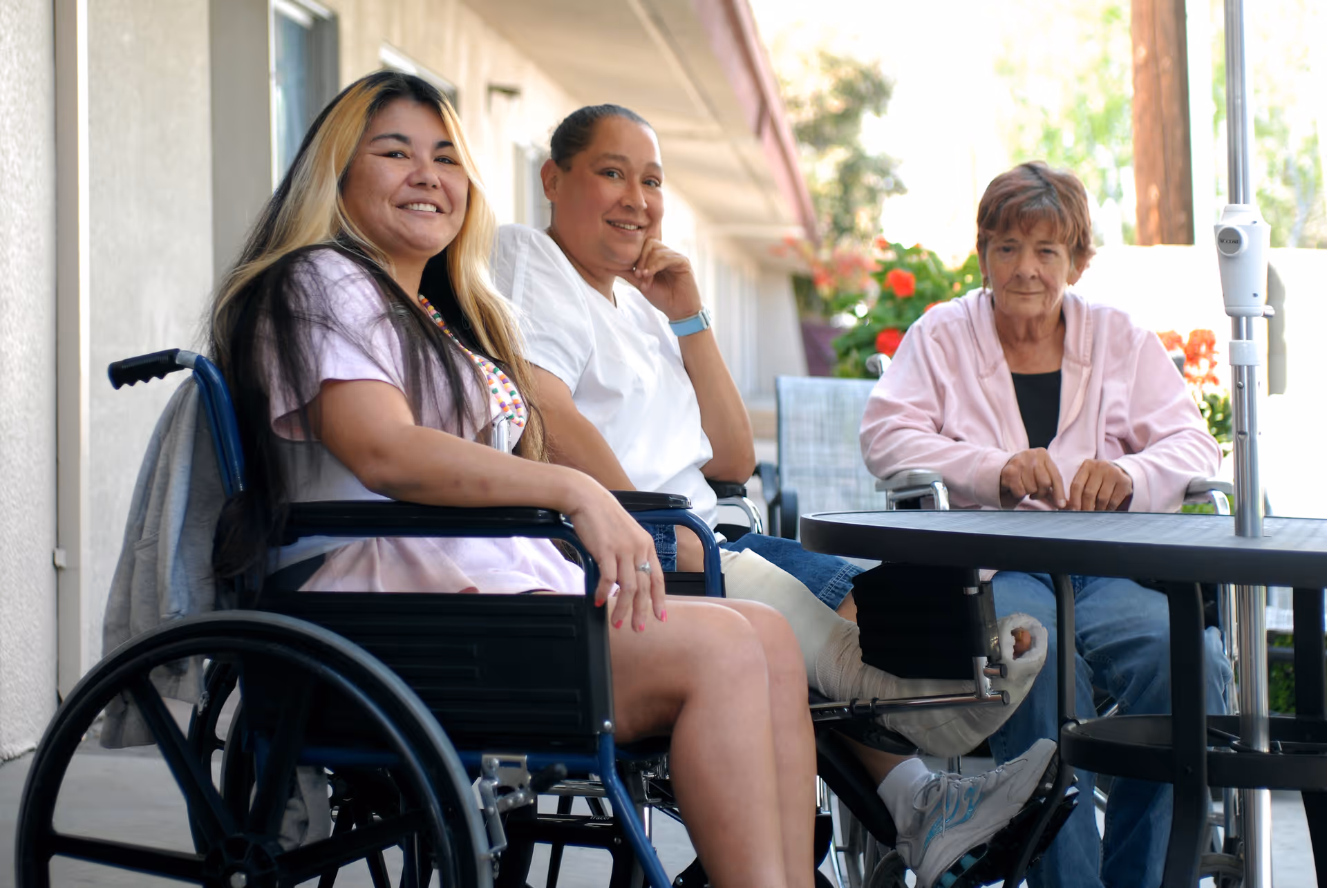 Three women sitting outside at a round table, two of them in wheelchairs. One woman has long hair and is smiling at the camera, the second woman is wearing a white shirt and has a cast on her leg, and the third woman is wearing a pink jacket and looking at the camera. They are on a patio area with plants and outdoor furniture.