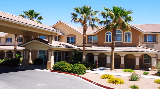 Exterior view of Parkway Assisted Living facility showing a two-story building with a tiled roof, multiple windows, palm trees, and landscaped bushes and plants in front of the entrance driveway.