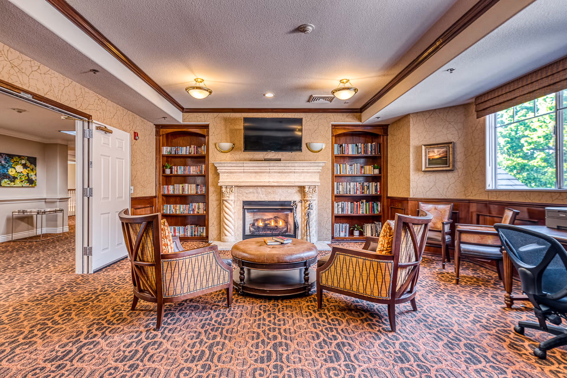 Cozy senior living common room with a lit fireplace, bookshelves, round ottoman and armchairs.