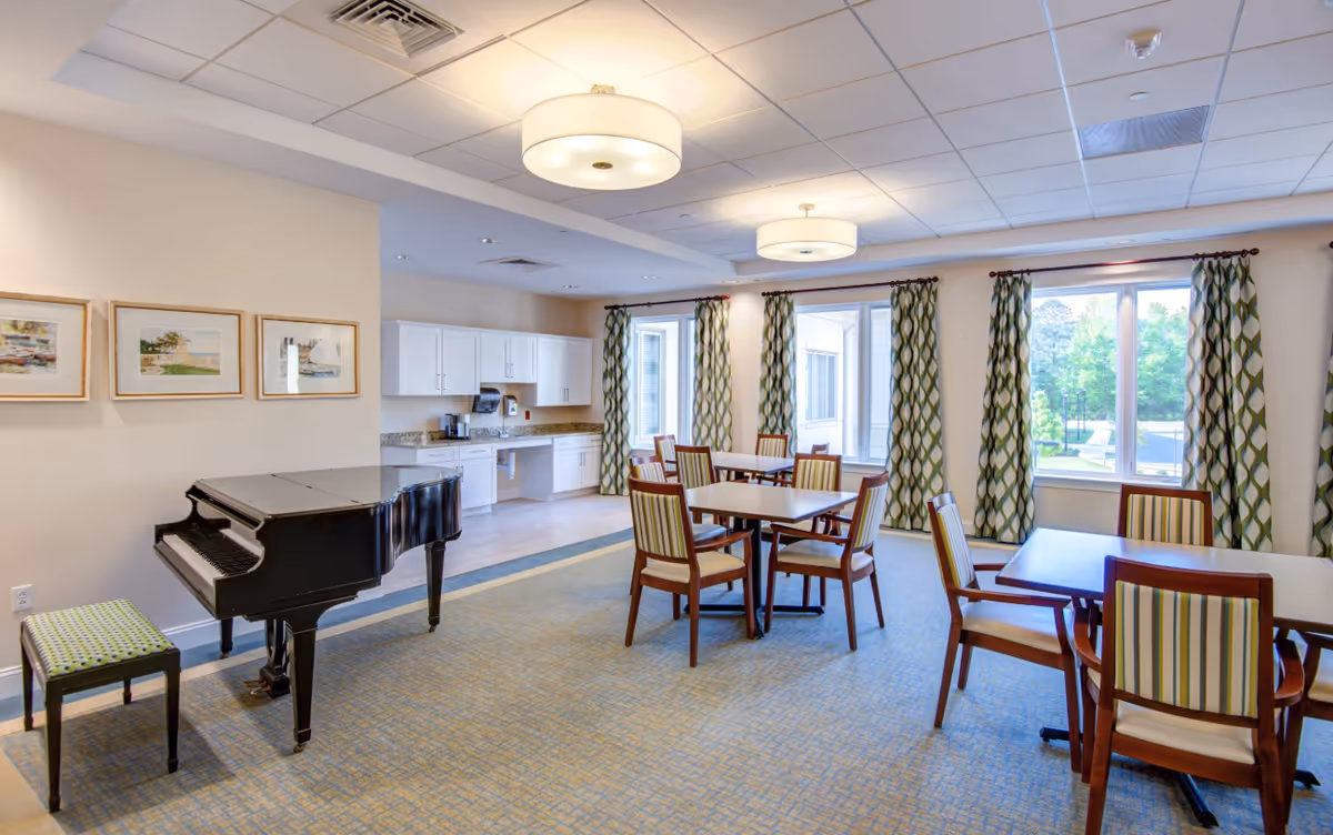 Bright communal dining room with tables and chairs, a grand piano, and a small kitchenette by large windows with patterned curtains.