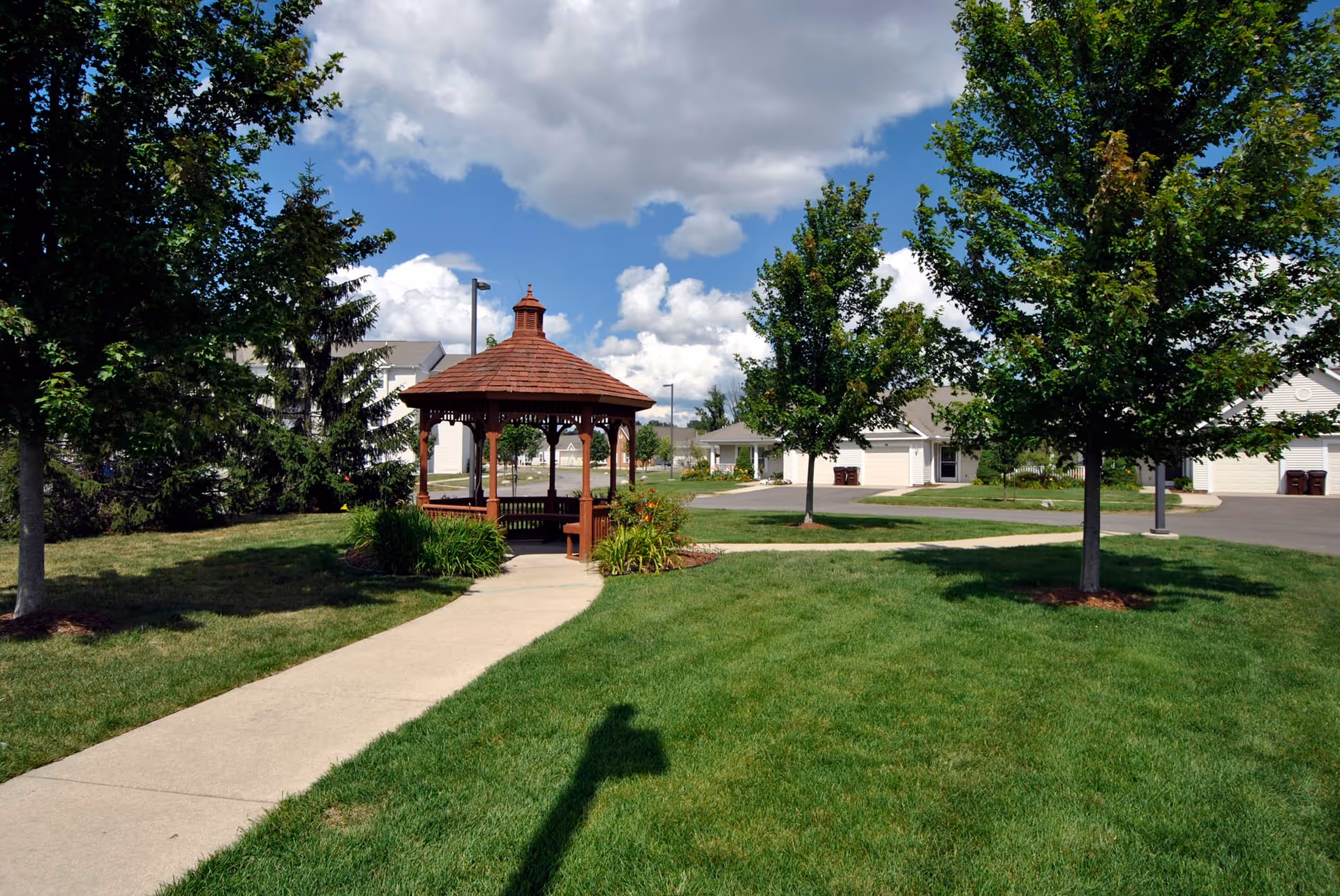 A well-maintained outdoor area at Grandhaven Manor featuring a wooden gazebo surrounded by green grass, trees, and a paved walkway under a partly cloudy sky.
