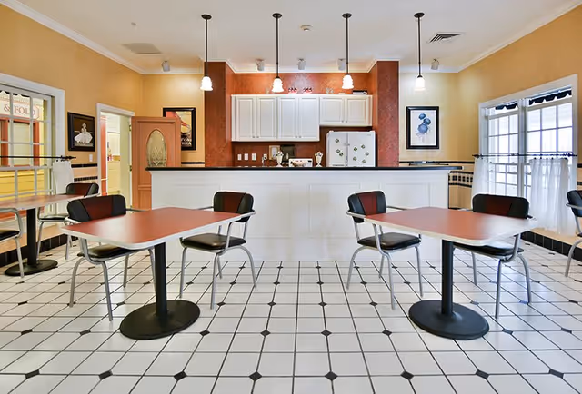 A bright dining area with white tiled floor featuring black diamond accents, several tables with red tops and black chairs, and a kitchen counter with white cabinets and pendant lights hanging above. The walls are painted yellow with framed artwork and windows with white curtains.