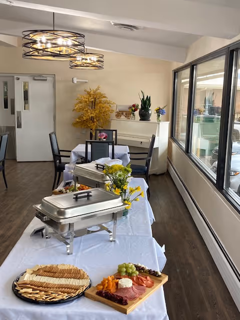 Buffet table with chafing dishes, a charcuterie board, and floral arrangements in a bright dining room with large windows and hanging lights.