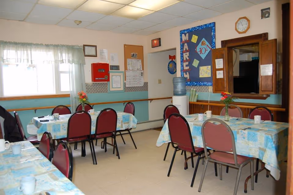 A dining room with several tables covered in blue and white patterned tablecloths. Each table has red cushioned chairs around it and a vase with flowers as a centerpiece. There is a water cooler against the wall, a bulletin board with notices, and a window with sheer curtains letting in natural light.