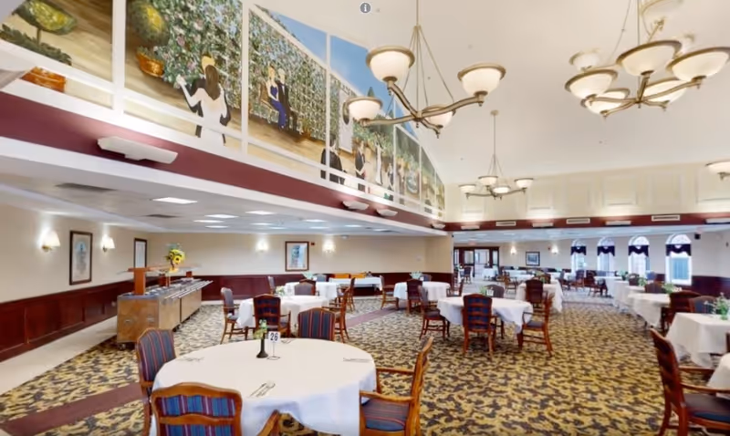 Large assisted-living dining room with round tables covered in white tablecloths, chandeliers, patterned carpet, and a mural on the upper wall.