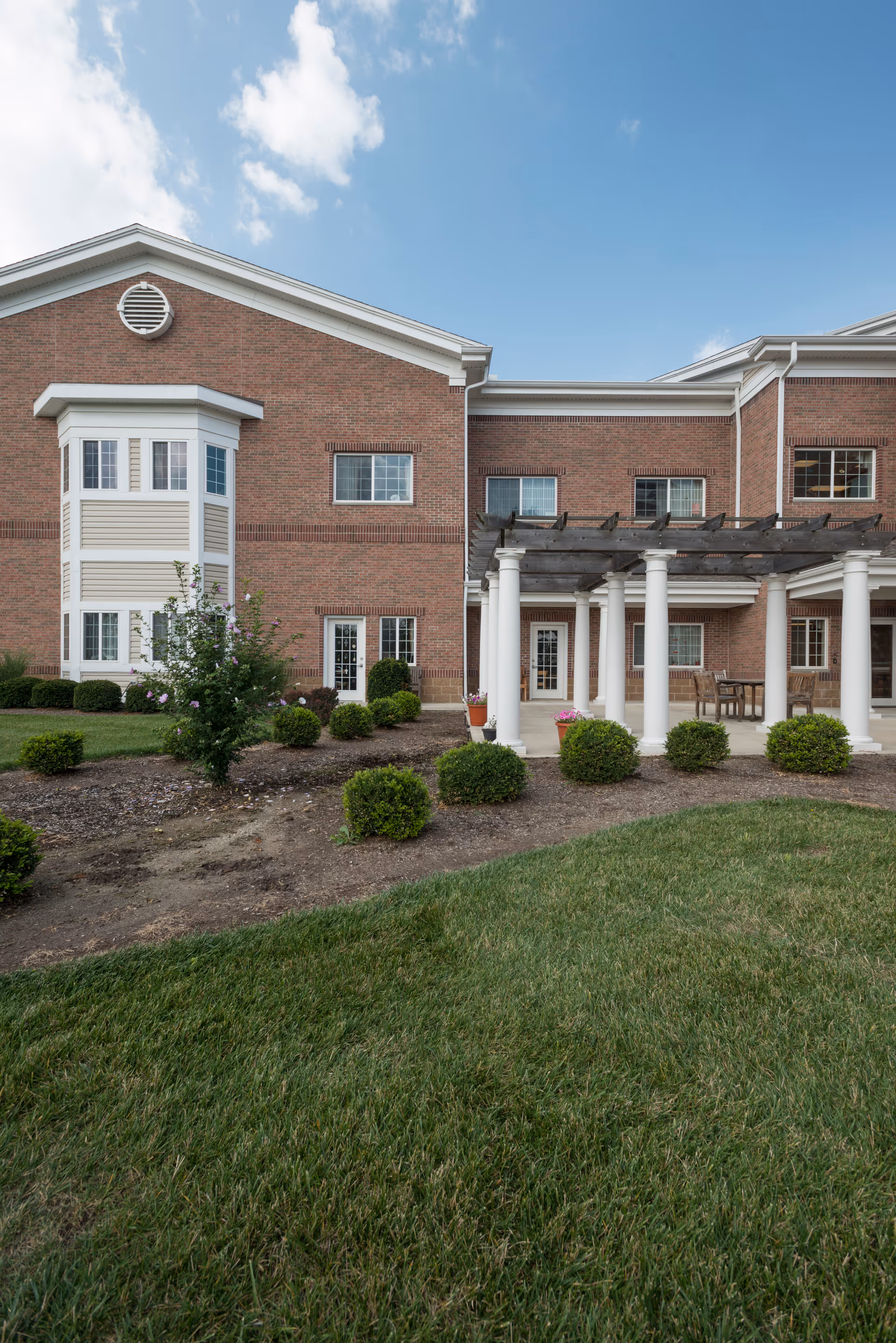 Brick senior living facility exterior showing white columns and a pergola, windows, and a landscaped lawn.