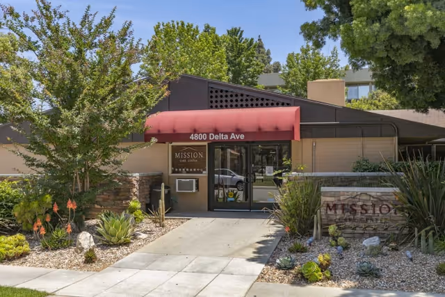 Entrance of the Mission Care Center building with a red awning reading "4800 Delta Ave", glass doors, and a landscaped walkway.