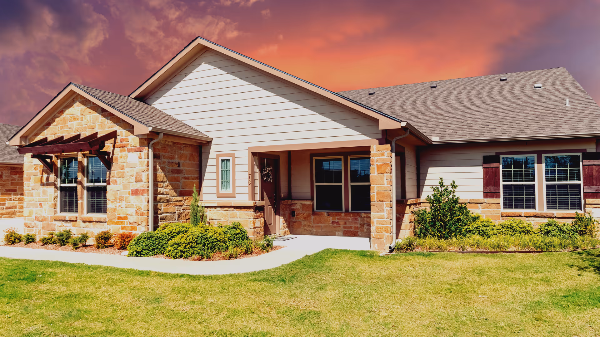 Exterior view of a single-story residential building with stone and siding facade, a small covered porch, windows with shutters, and landscaped greenery in front under a vibrant orange and purple sky at sunset.