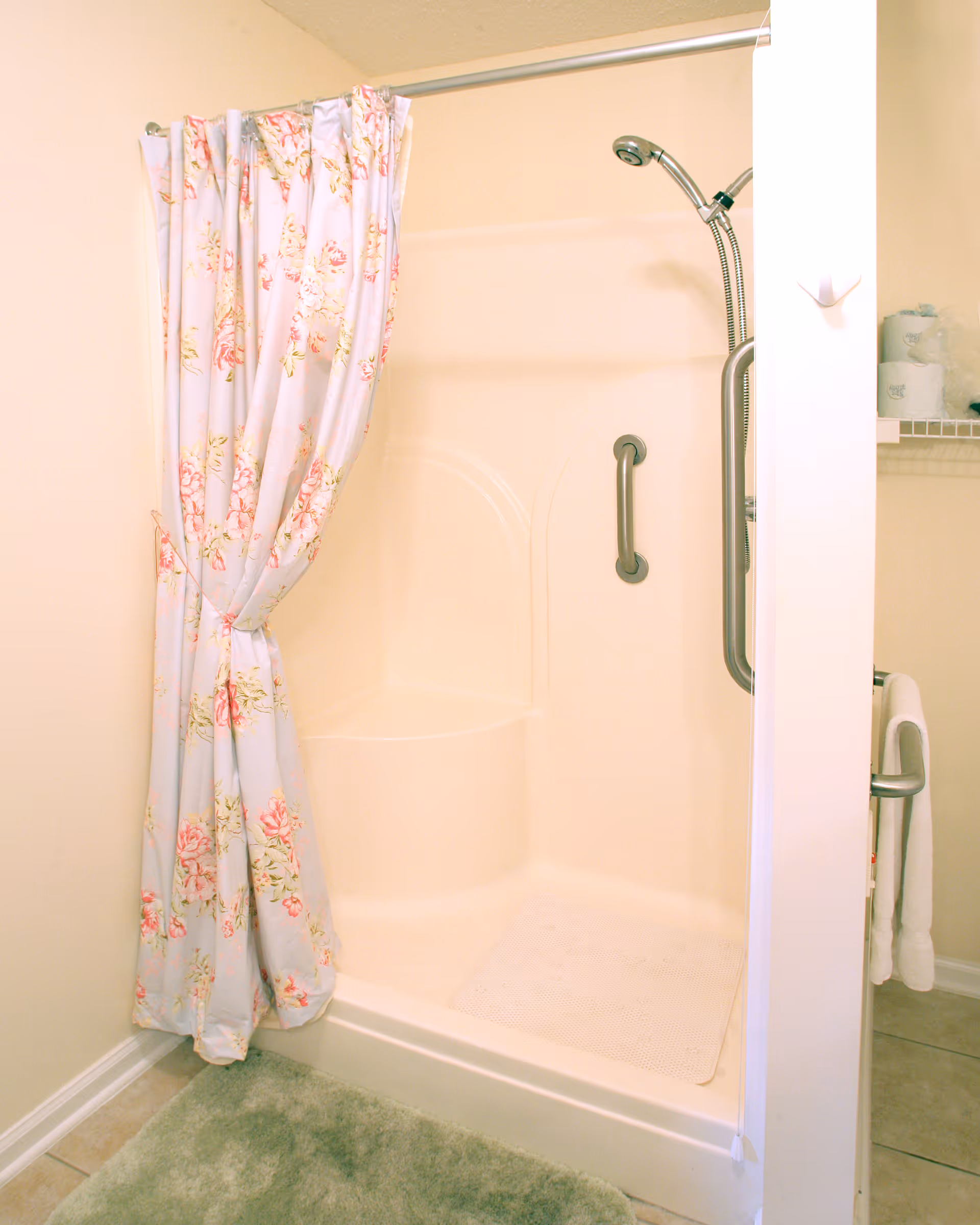 A bathroom shower area with a floral shower curtain tied to one side, a built-in seat, grab bars for safety, a handheld showerhead, and a green bath mat on the floor outside the shower.