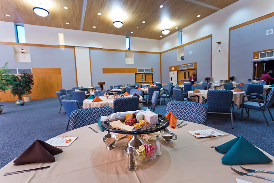A spacious dining room with multiple round tables covered with beige tablecloths, each set with folded napkins in various colors, menus, and silverware. The room has blue patterned chairs, a high wooden ceiling with circular lights, and large windows near the ceiling. A centerpiece with cheese, crackers, and grapes is on the closest table.