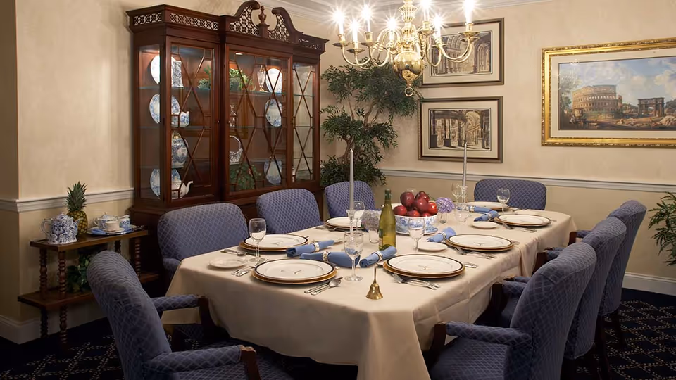 A formal dining room with a rectangular table covered with a beige tablecloth, set with plates, wine glasses, silverware, and blue napkins. The table is surrounded by eight upholstered chairs with a blue diamond pattern. A wooden china cabinet with glass doors displays blue and white china. The room is decorated with framed artwork, a chandelier, and potted plants.