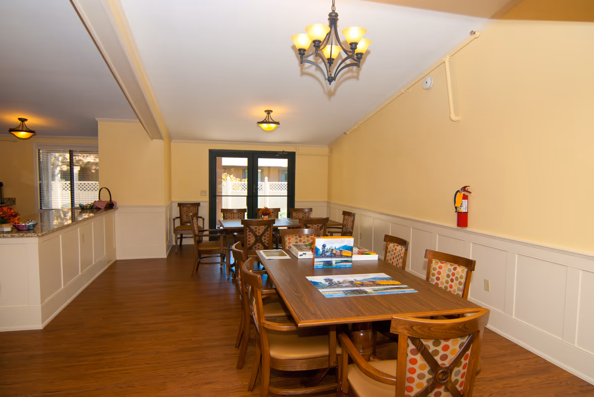 A well-lit interior room with wooden flooring and cream-colored walls. The room features a long wooden table surrounded by chairs with patterned cushions. On the table, there are puzzles and books. In the background, there is another smaller table with chairs near glass doors leading outside. Ceiling lights hang from the ceiling, and a fire extinguisher is mounted on the wall.