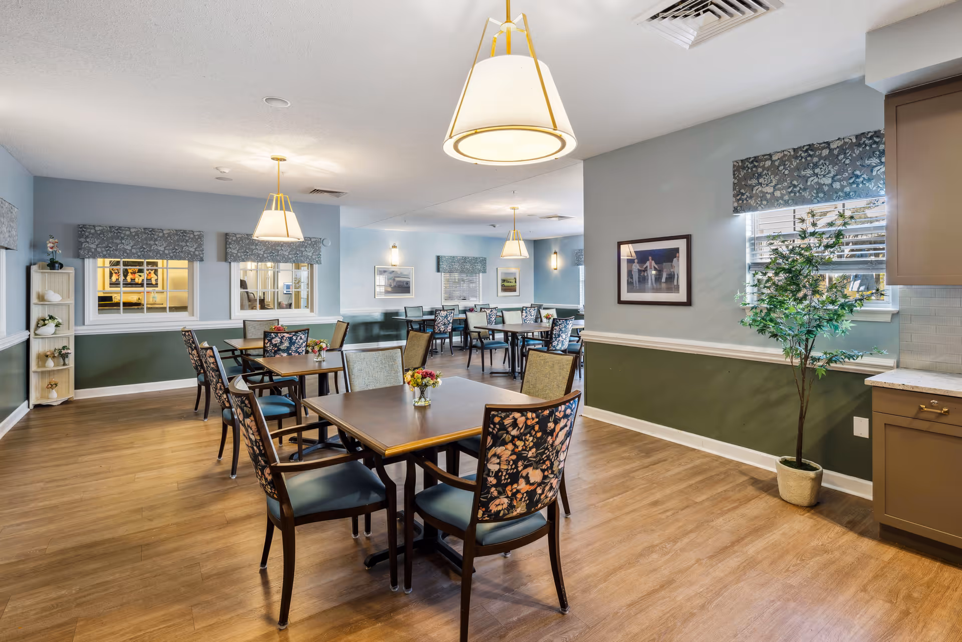 A well-lit dining room with several wooden tables and chairs, some with floral upholstery. The room has light blue and green walls, wooden flooring, and large pendant lights hanging from the ceiling. There are windows with floral valances, framed pictures on the walls, and a potted plant near a window.