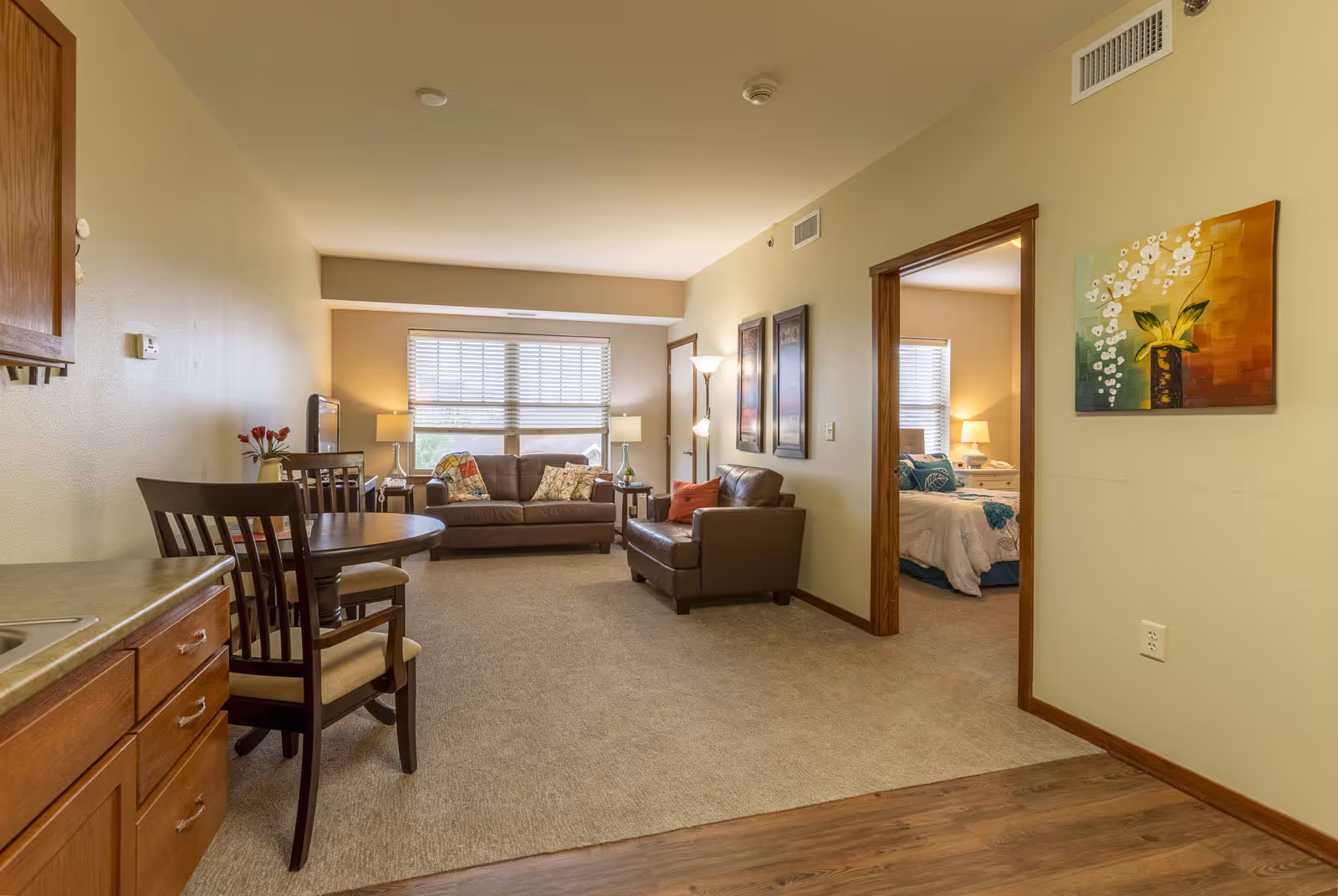 Interior view of a senior living apartment showing a living room with two brown leather sofas, a TV on a stand, and a window with blinds. To the left is a small dining area with a round wooden table and chairs, and part of a kitchen counter with cabinets is visible. To the right, an open doorway leads to a bedroom with a bed, nightstand, and lamp. The walls are painted light beige and decorated with framed artwork and a floral painting.