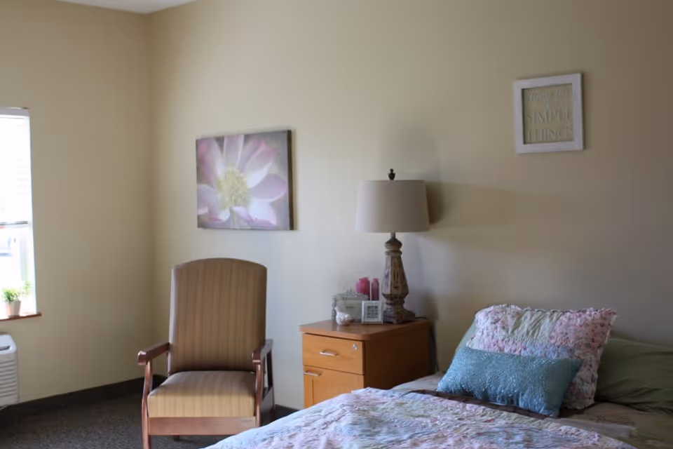 A cozy bedroom with a bed covered in floral bedding and multiple pillows, a wooden nightstand with a decorative lamp and candles, a beige upholstered armchair, and two framed pictures on the light-colored walls.