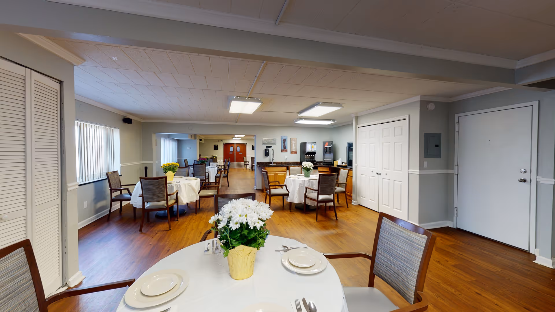 A dining room in Imperial Senior Suites featuring several round tables covered with white tablecloths, each set with plates, utensils, and a flower centerpiece. The room has wooden flooring, light gray walls, and windows with vertical blinds. Chairs with wooden frames and cushioned seats surround the tables. In the background, there is a beverage station with coffee machines and a phone mounted on the wall.