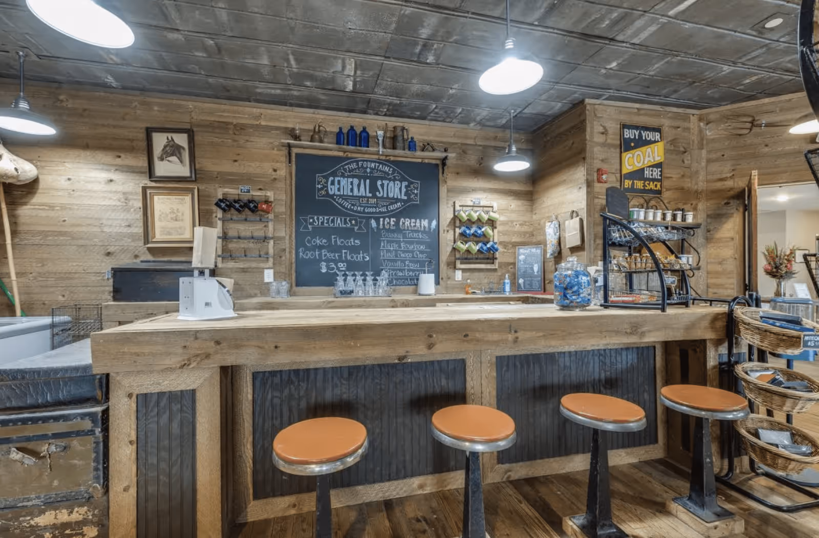 Interior view of a rustic general store setup with a wooden counter and five orange cushioned stools. The walls are wooden with various decorations including framed pictures, a chalkboard menu listing specials like Coke floats and root beer floats, and shelves with bottles and items. Overhead industrial-style lights illuminate the space.