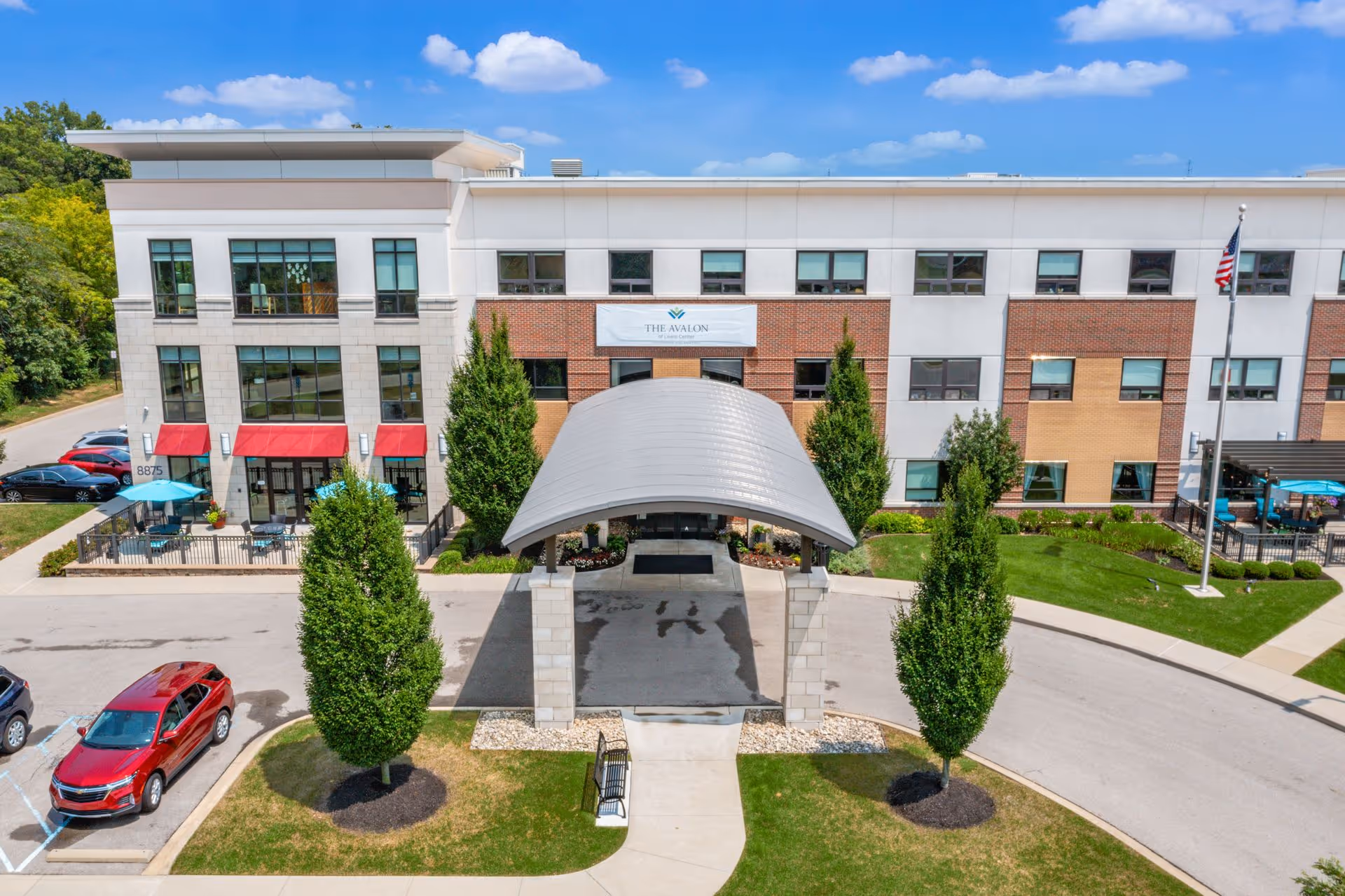 Front exterior view of The Avalon of Lewis Center building with a covered entrance, three tall trees, a flagpole with an American flag, parked cars, and outdoor seating areas with blue umbrellas on a sunny day.