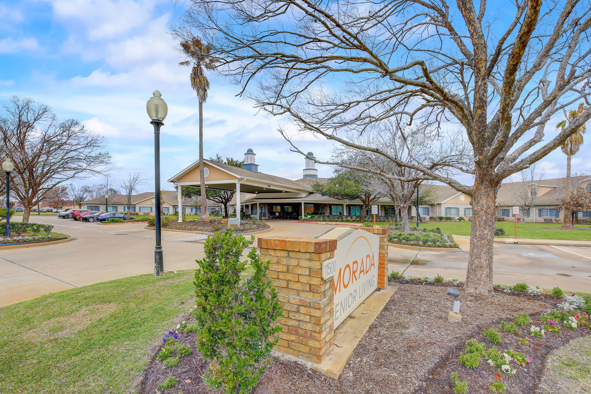 Exterior view of Morada Senior Living facility showing the entrance with a covered driveway, a brick sign with the facility name, landscaped greenery, trees without leaves, and a parking area with several cars under a partly cloudy sky.