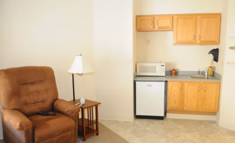 A small living area with a brown recliner chair next to a wooden side table with a white lamp. Adjacent to the chair is a kitchenette with wooden cabinets, a small refrigerator, a microwave, a sink, and a soap dispenser mounted on the wall.