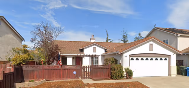 Single-story home with a red wooden front fence and gate, an attached two-car garage, small front yard and American flag under a blue sky.