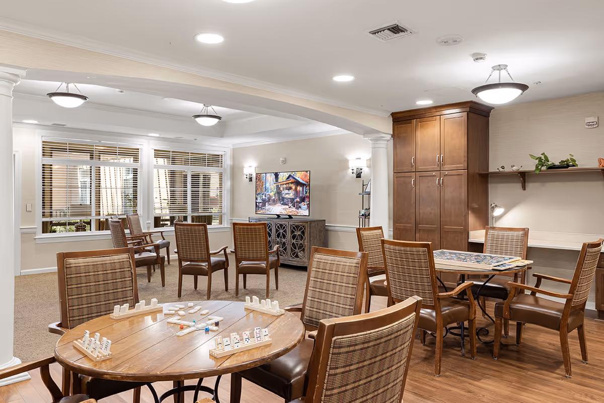 A well-lit common area in a senior living facility with multiple wooden tables and chairs arranged for games and social activities. One table has a domino game set up, and another table has a puzzle in progress. The room features beige walls, a large window with blinds, a TV mounted on a cabinet, and wooden cabinetry with shelves holding decorative items and plants.