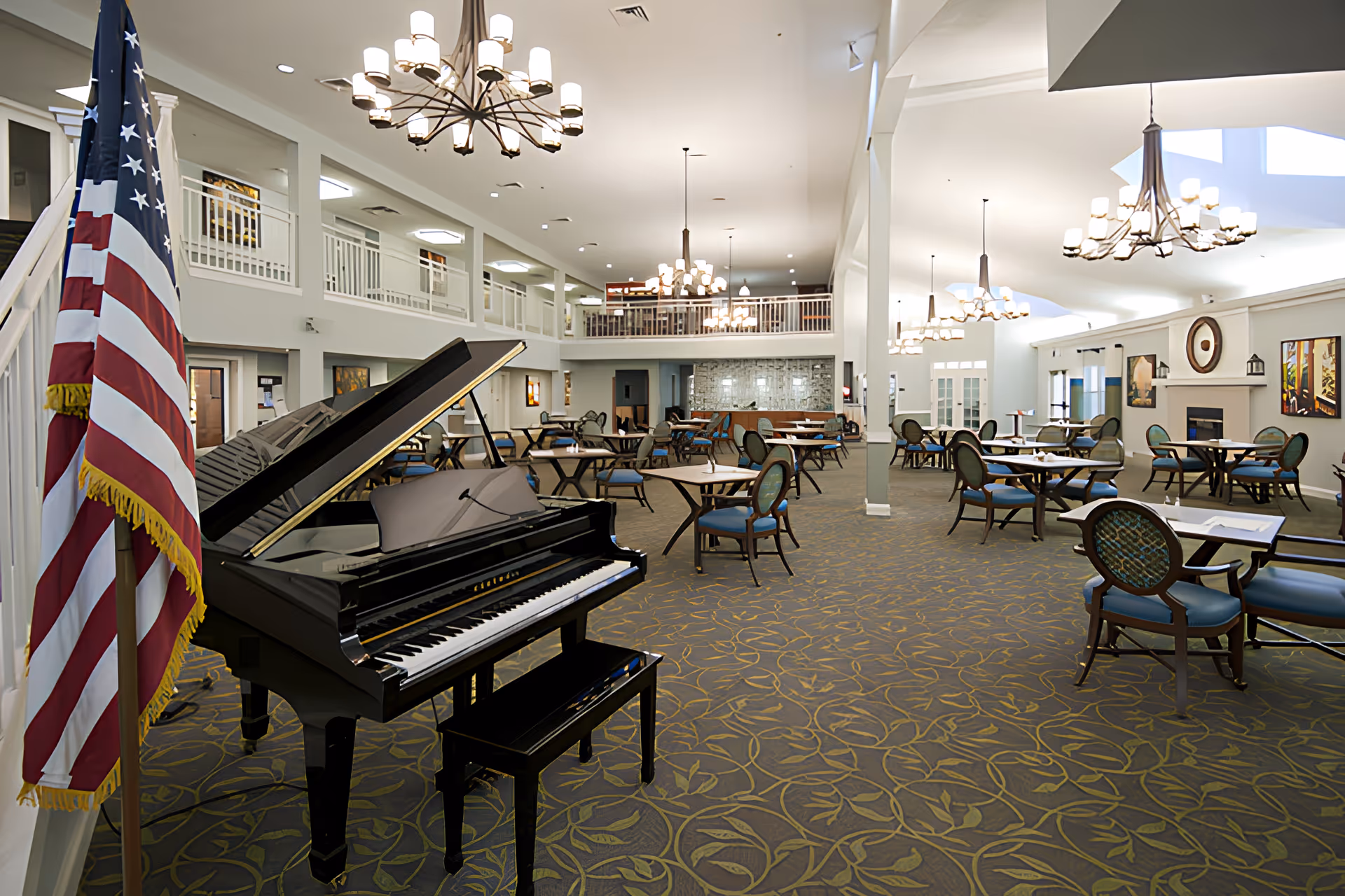 Spacious senior living facility common area with a grand piano and American flag in the foreground. The room features multiple round tables with chairs, elegant chandeliers, a fireplace, and a second-floor balcony overlooking the area.