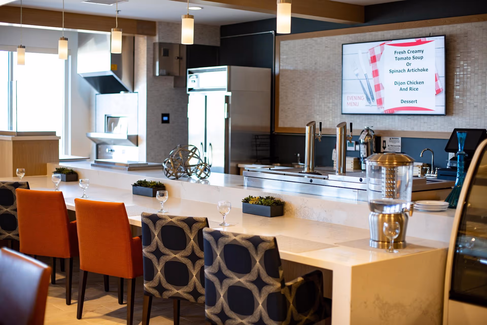 A modern dining area with a long white counter and a mix of orange and patterned chairs. There are small plants and glasses on the counter. Behind the counter is a kitchen area with stainless steel appliances, a water dispenser, and a digital menu board displaying the evening menu. Pendant lights hang from the ceiling, providing warm lighting.