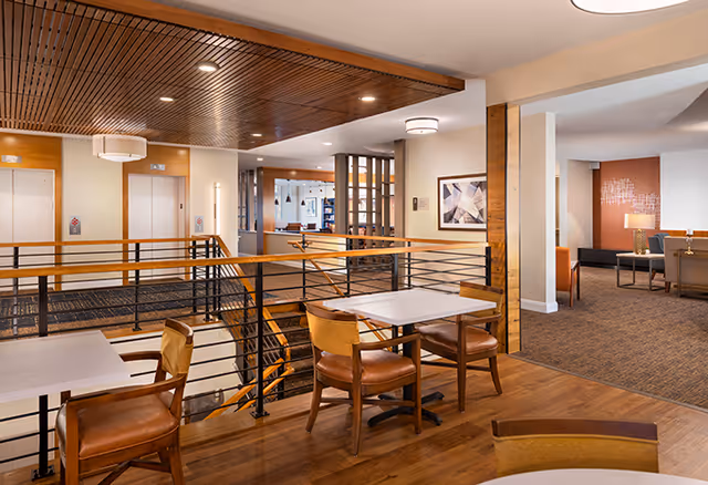 Interior view of a senior living facility showing a seating area with tables and chairs near a railing overlooking a lower level. The space features wooden flooring, a wooden slatted ceiling, two elevators in the background, and a lounge area with sofas and lamps visible through an open doorway.