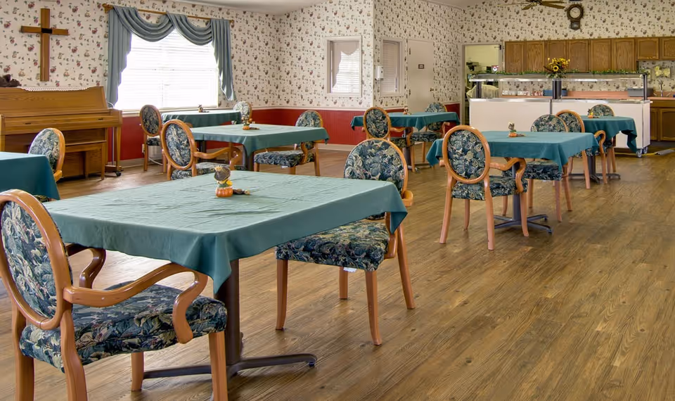A dining room with several tables covered in teal tablecloths and floral upholstered chairs. The room has wood flooring, floral wallpaper, a window with blue curtains, a wooden piano, and a buffet area with cabinets and a clock on the wall.